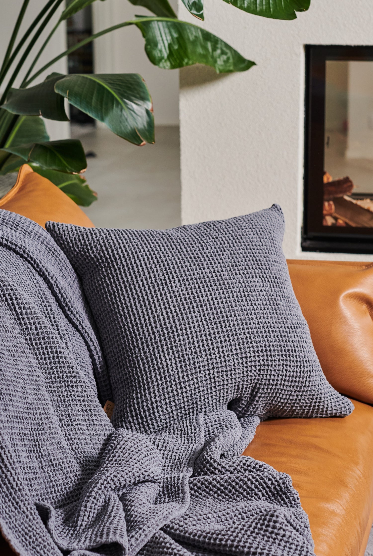 Gray textured pillow on a brown leather couch with a plant and fireplace in the background