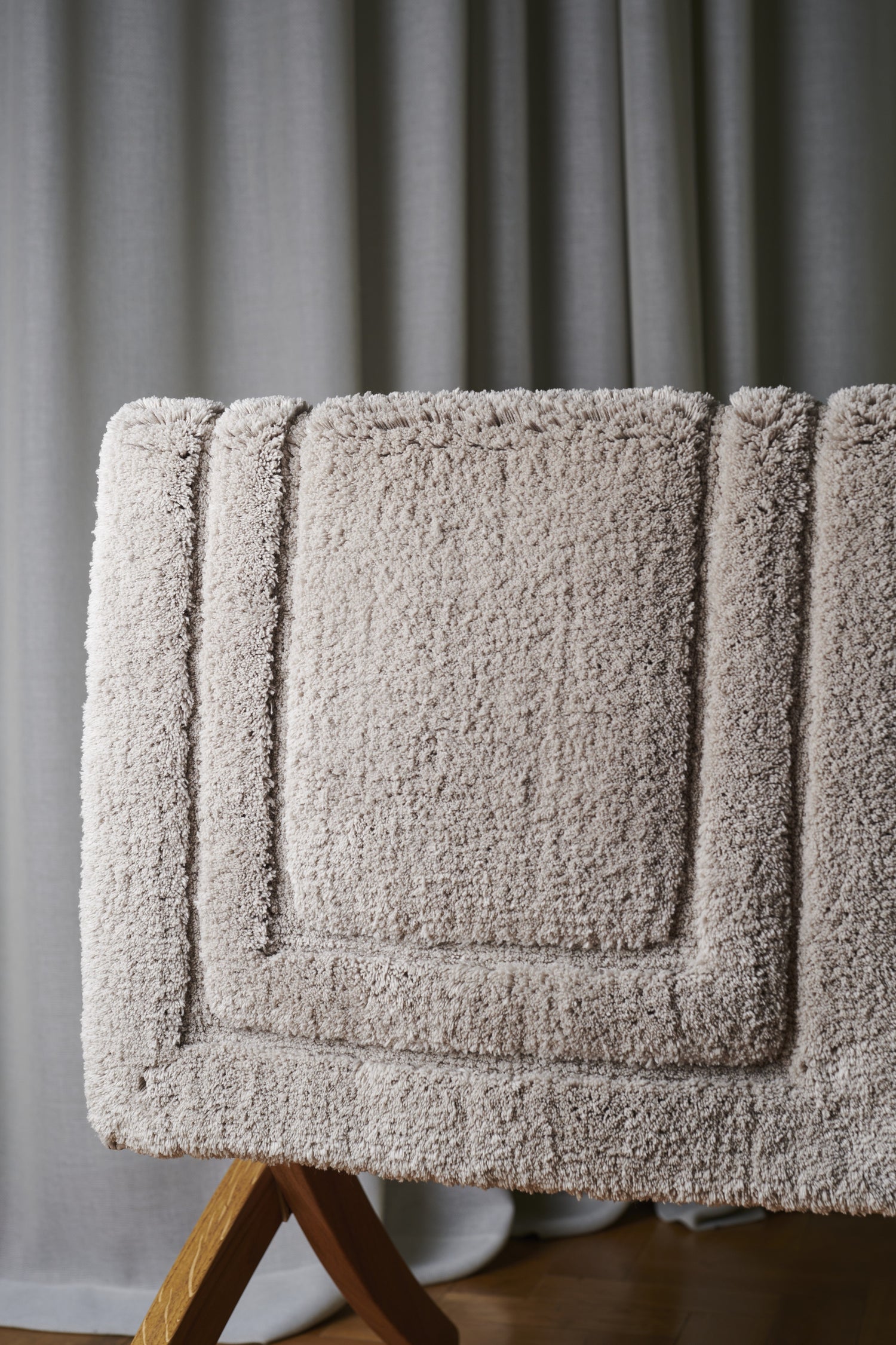 Textured beige bath mat on a wooden stand against a gray curtain background