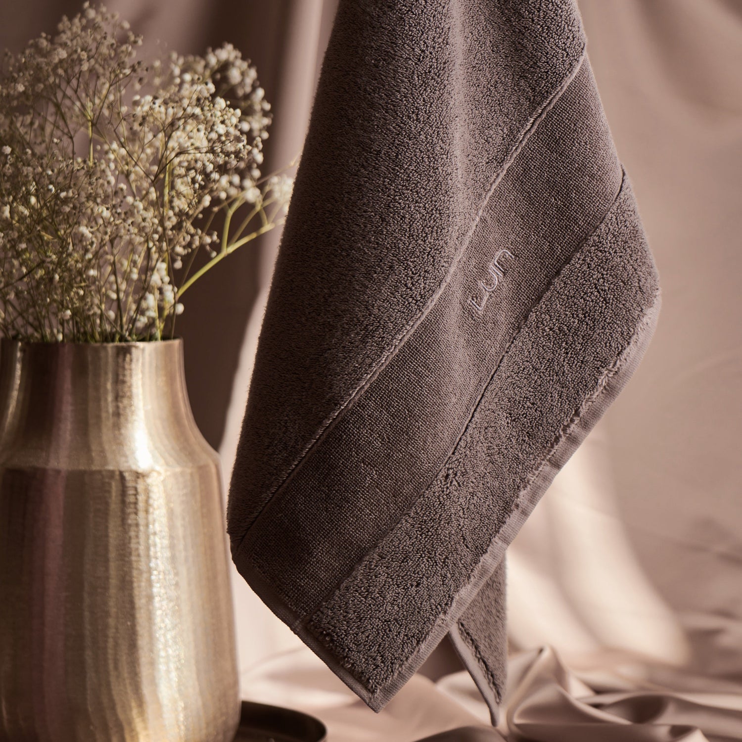 Gray towel with a brand logo draped over a neutral background with a vase of flowers.