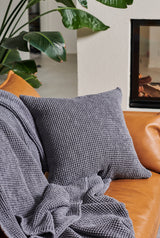 Gray textured pillow on a brown leather couch with a plant and fireplace in the background