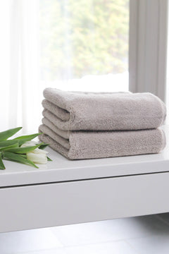 Two folded beige cotton towels stacked on a white vanity table next to white tulips, with a softly blurred window background.